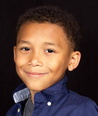 Young boy with short curly hair smiling confidently, wearing a navy blue shirt, against a dark background.