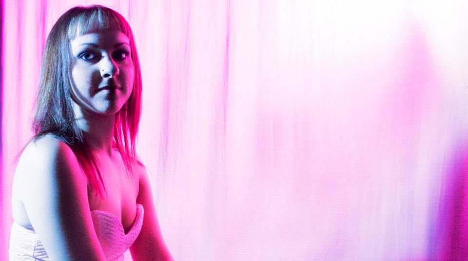 Young woman with blonde hair posing in pink and purple neon lighting against a soft pink curtain backdrop.