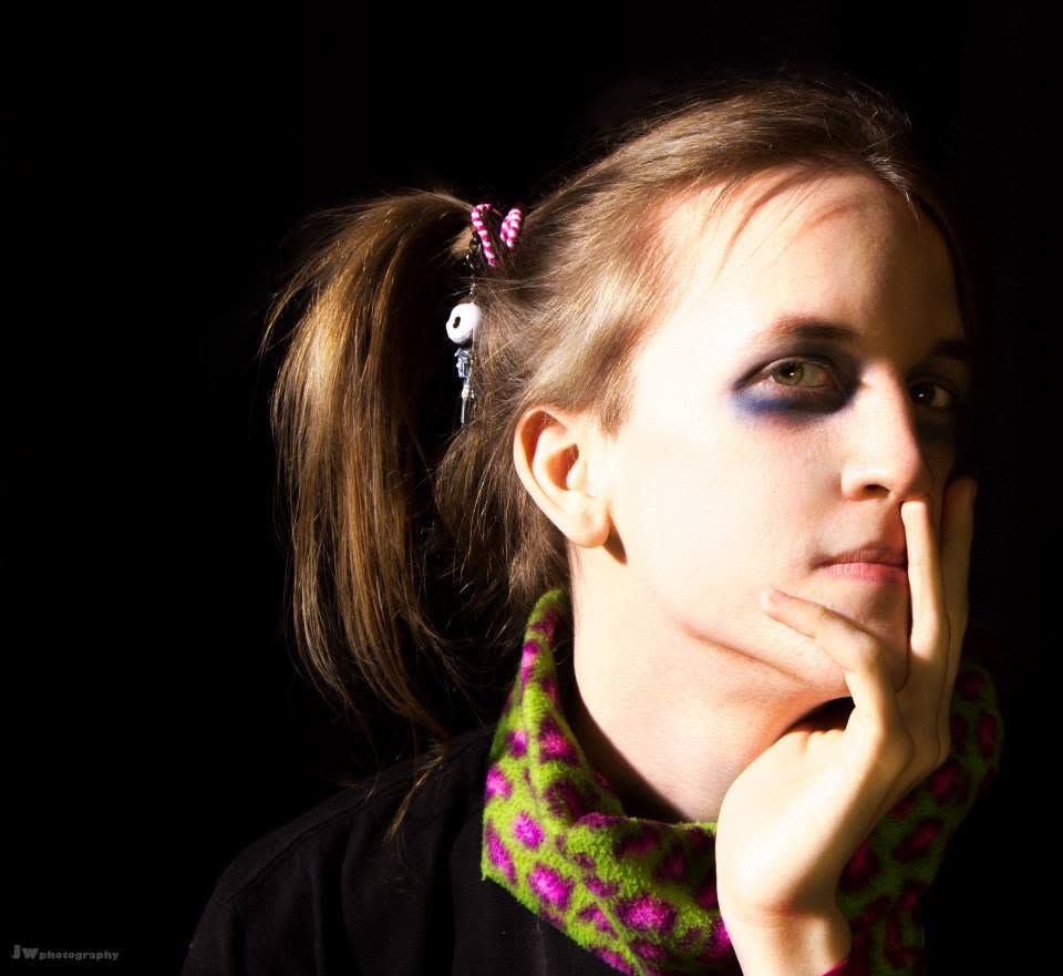 Young woman with ponytail and dramatic eye makeup, resting chin on hand against a dark background.