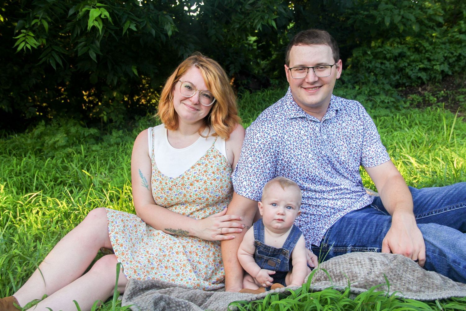 A young family with a baby sits on a blanket in a lush green park, smiling warmly for a portrait photo.