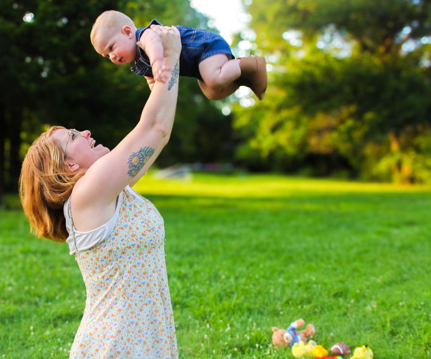 A joyful mother lifts her baby boy high in the air in a lush green park on a sunny day.