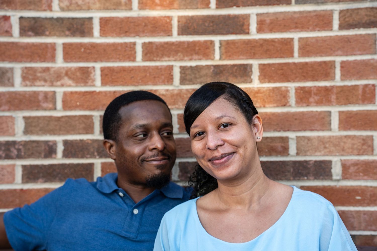 A smiling couple poses together against a brick wall, the man in a blue shirt and woman in a light blue top.