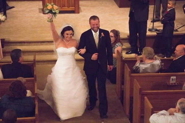 Bride raising bouquet while walking down church aisle with groom after wedding ceremony.