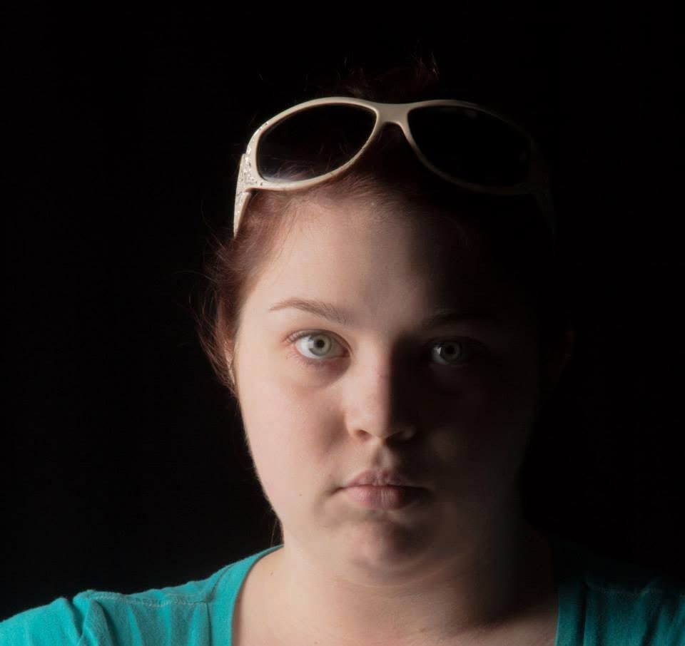 Young woman with white sunglasses on her head wearing a teal shirt against a dark background.