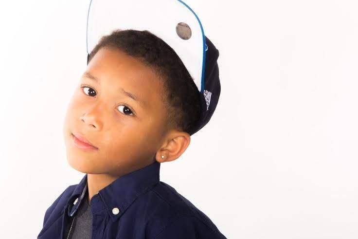 Young boy wearing a backwards blue cap and navy shirt, looking over his shoulder against white background.