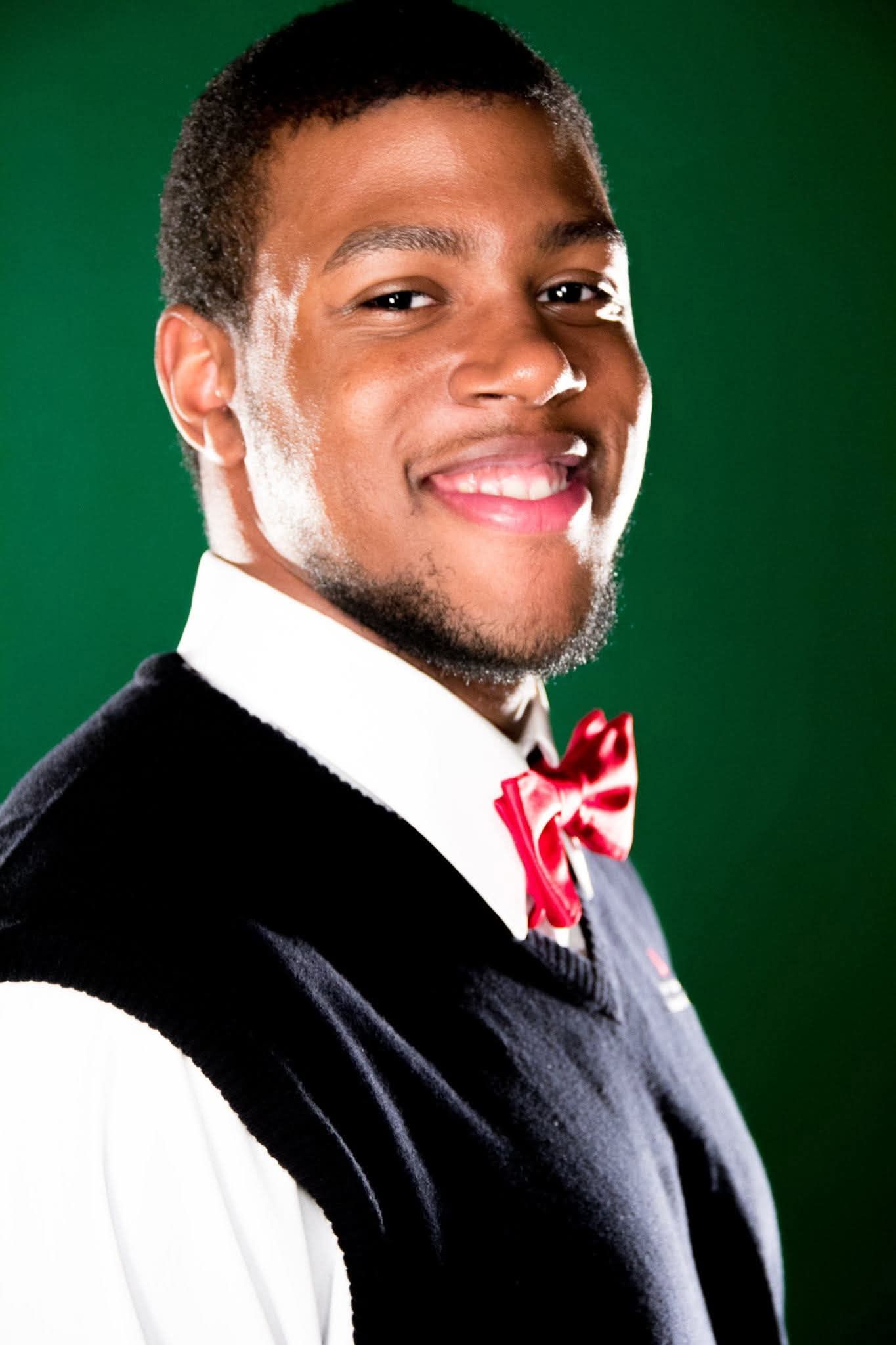 Smiling young man wearing a red bow tie, white shirt, and black vest against a green background.