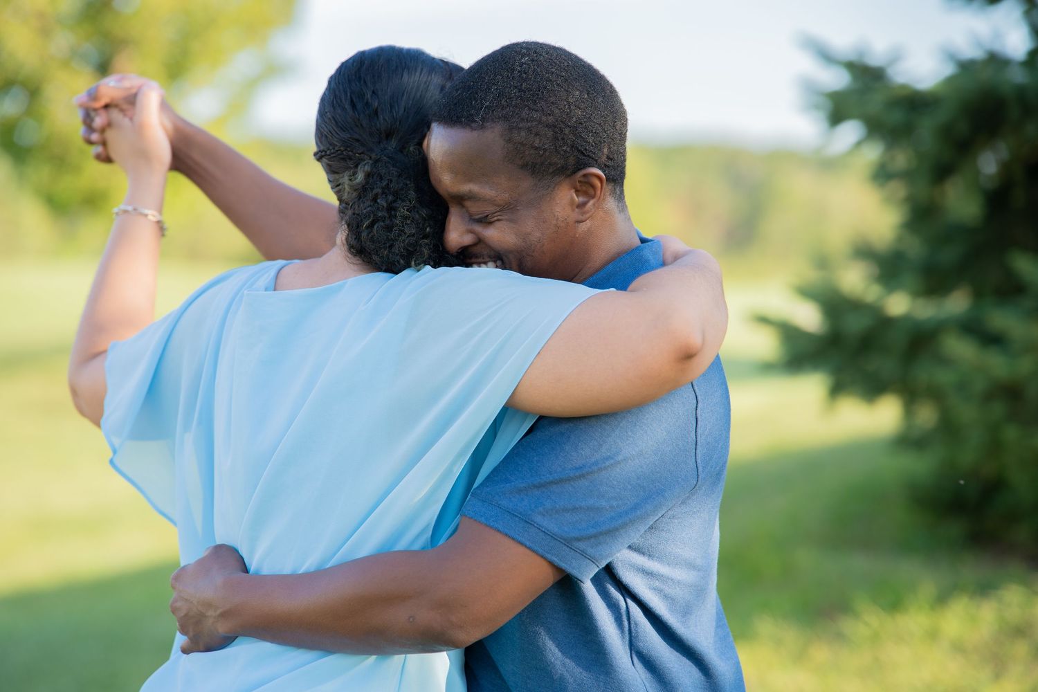 A couple embraces warmly outdoors, holding hands while hugging in a lush green park setting.