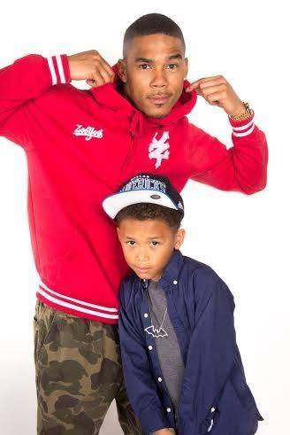 Man in red hoodie and camo pants posing with young boy in blue shirt and snapback cap.