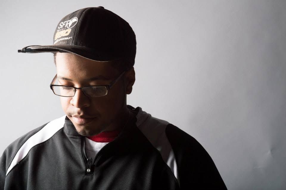 Man wearing glasses and black athletic jacket with snapback cap looks downward in moody portrait.