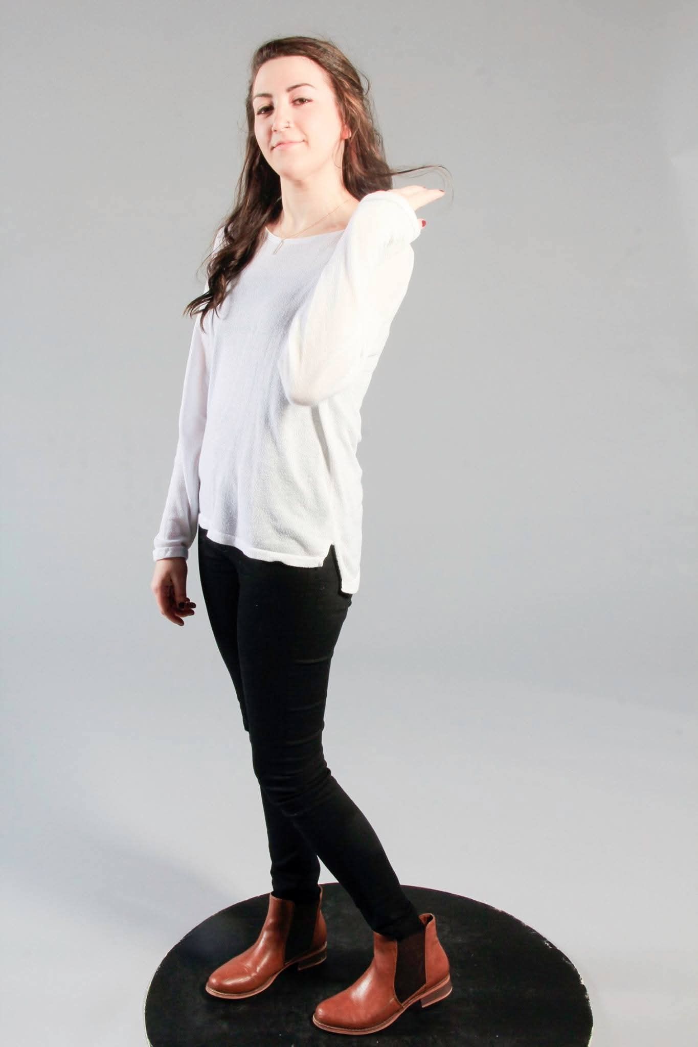 Young woman in white top, black skinny jeans, and brown ankle boots standing on a platform against gray background.