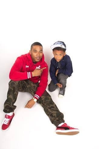 Man in red hoodie and camo pants sitting on floor pointing at young boy in blue shirt and cap.