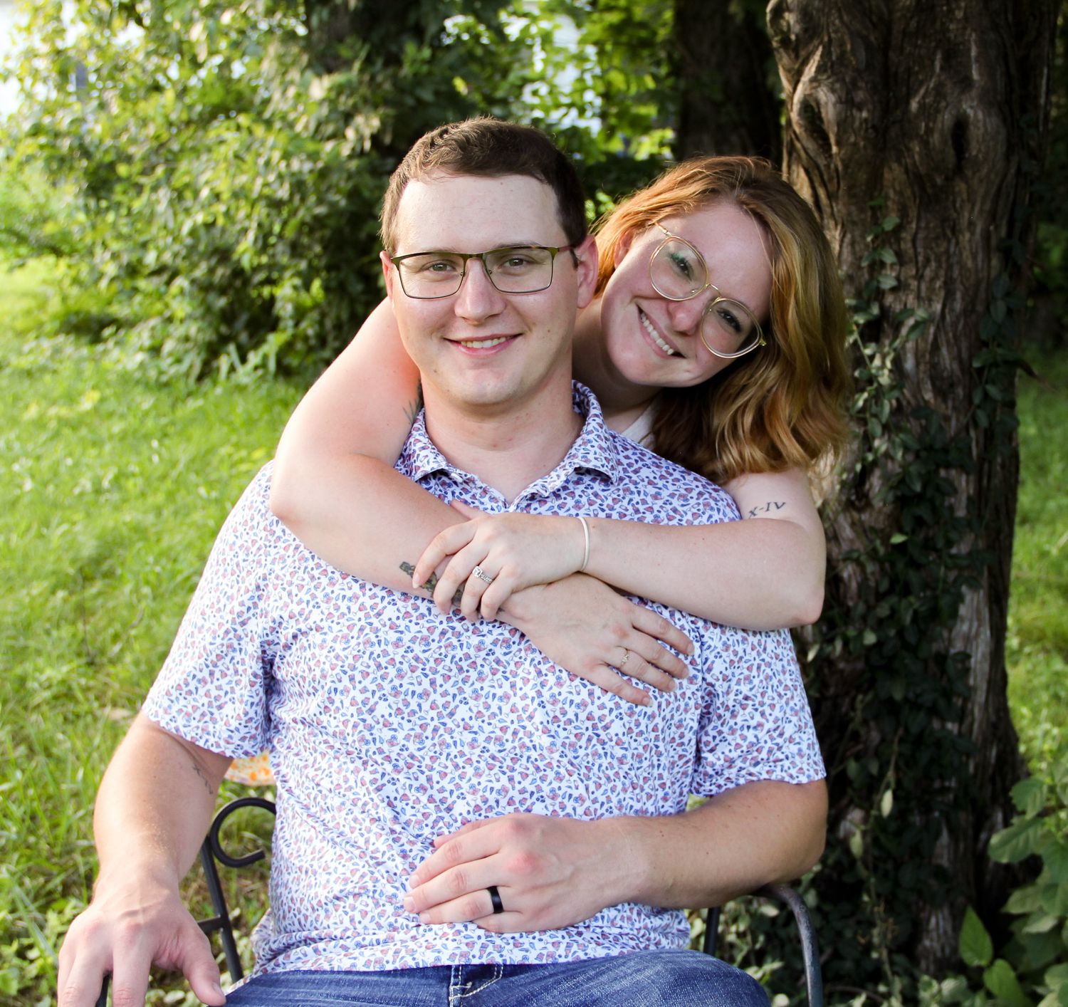 A smiling couple poses outdoors near a tree, woman hugging man from behind in a sunny green garden setting.