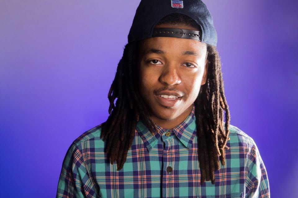 Smiling teenage boy with dreadlocks wearing a plaid shirt and navy cap against a purple background.