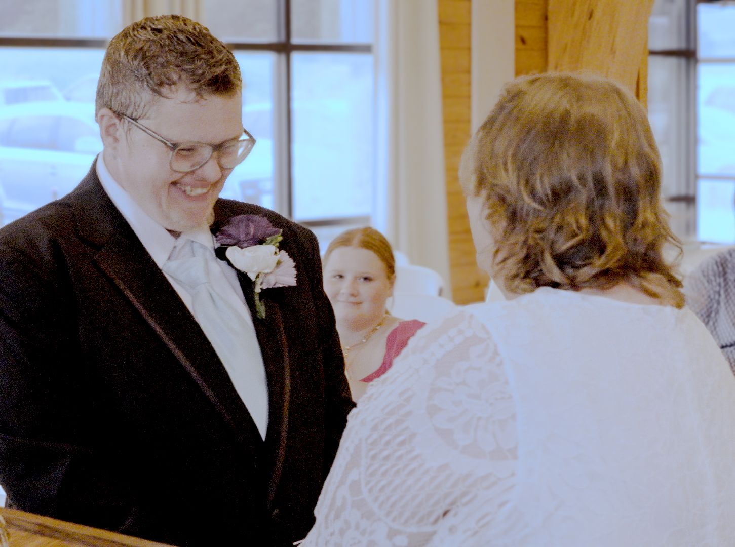 Groom in tuxedo smiling during wedding ceremony with bridesmaid and officiant in background.