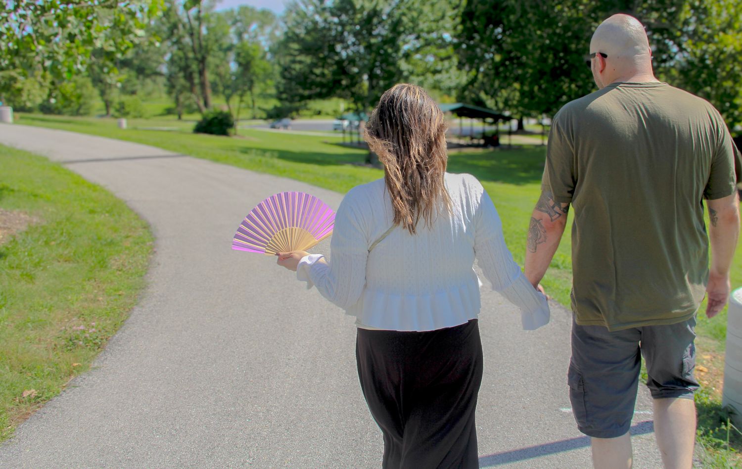 Couple walking hand in hand on a park path, woman holding a pink decorative fan on a sunny day.