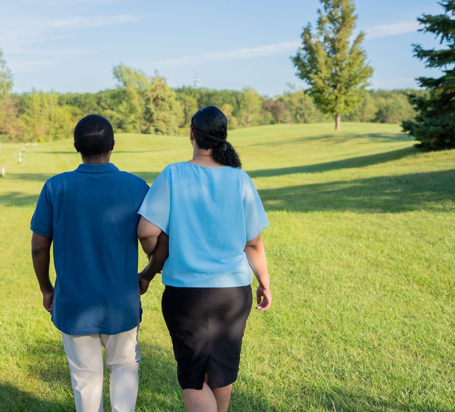 Two people walking arm in arm across a lush green golf course surrounded by trees on a sunny day.