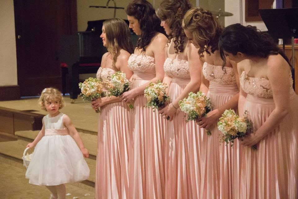 Bridesmaids in pink gowns holding white bouquets stand in a row as a little flower girl in white dress walks beside them.