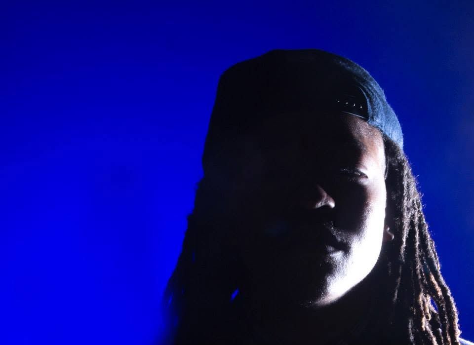 Person with dreadlocks wearing a cap, dramatically lit against a vivid blue background.