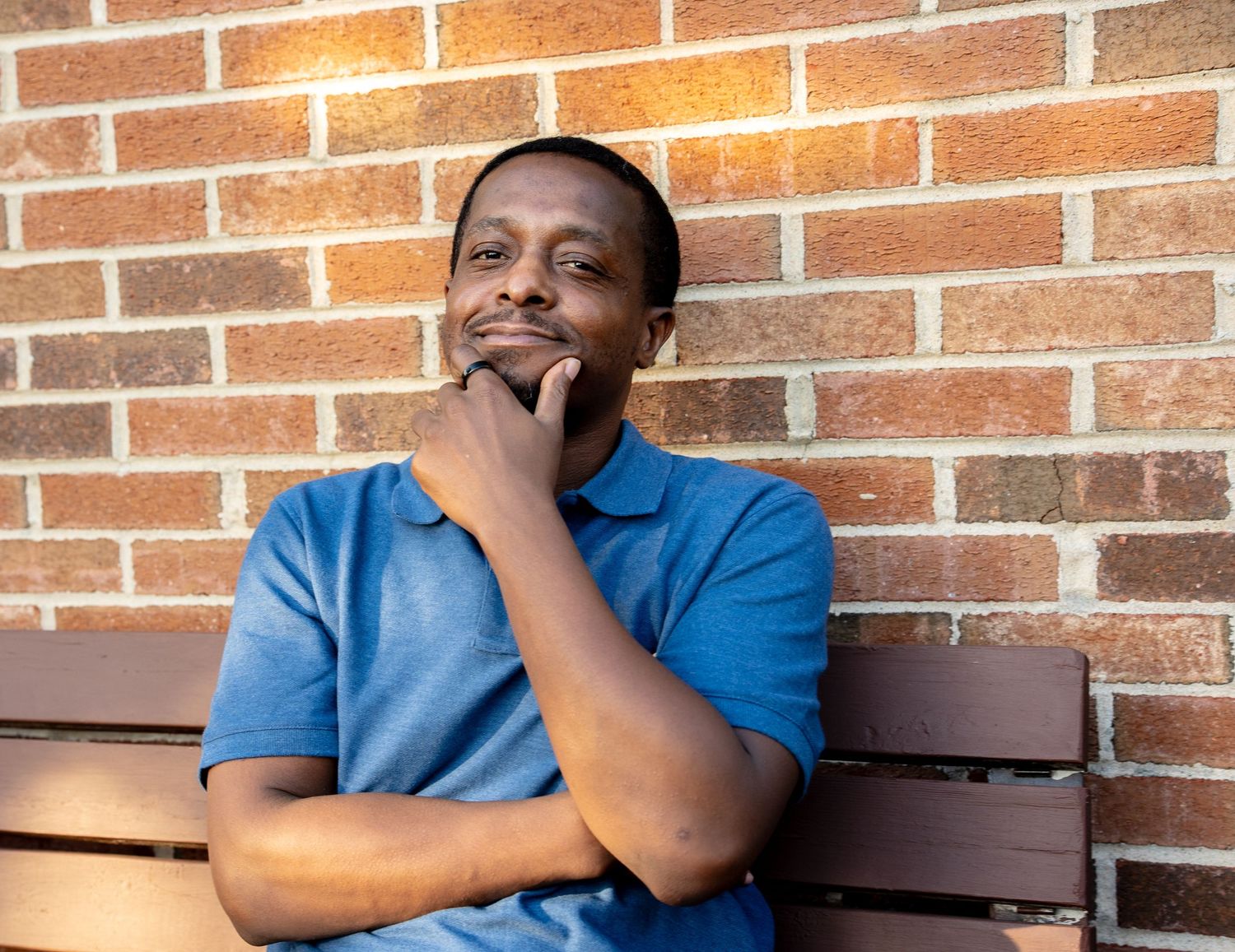 Thoughtful man in blue polo shirt sitting on bench against brick wall with hand on chin.