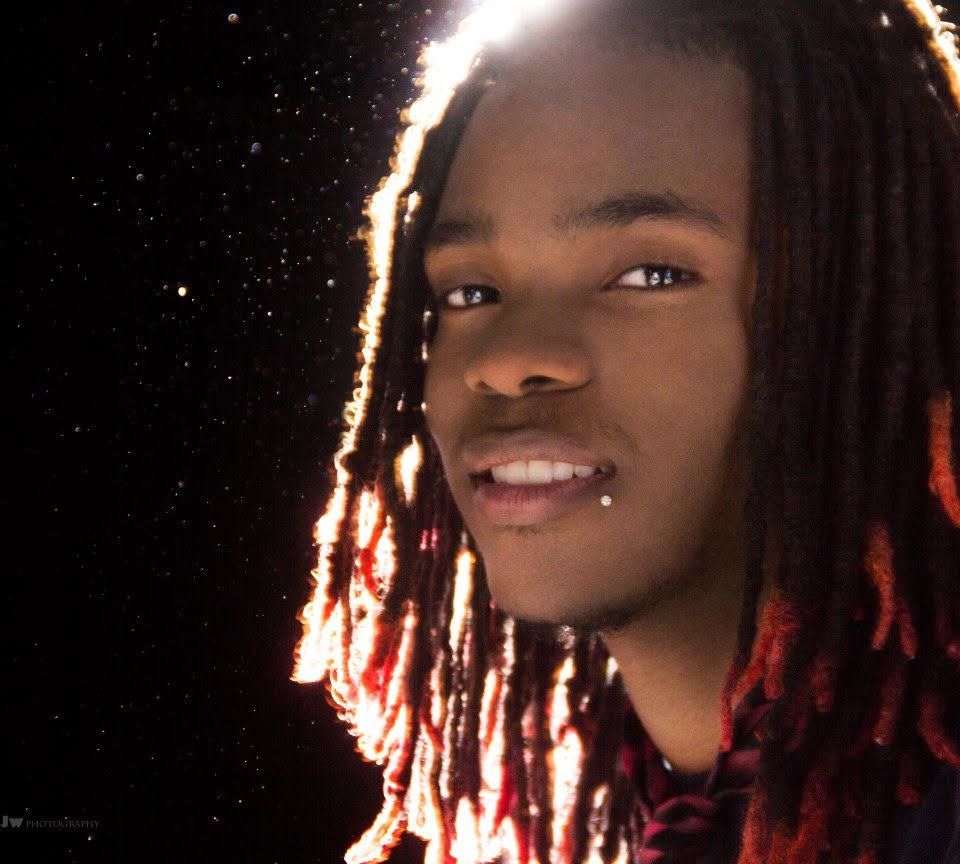 Young man with red-tipped dreadlocks and a chin piercing smiling against a dark starry background.