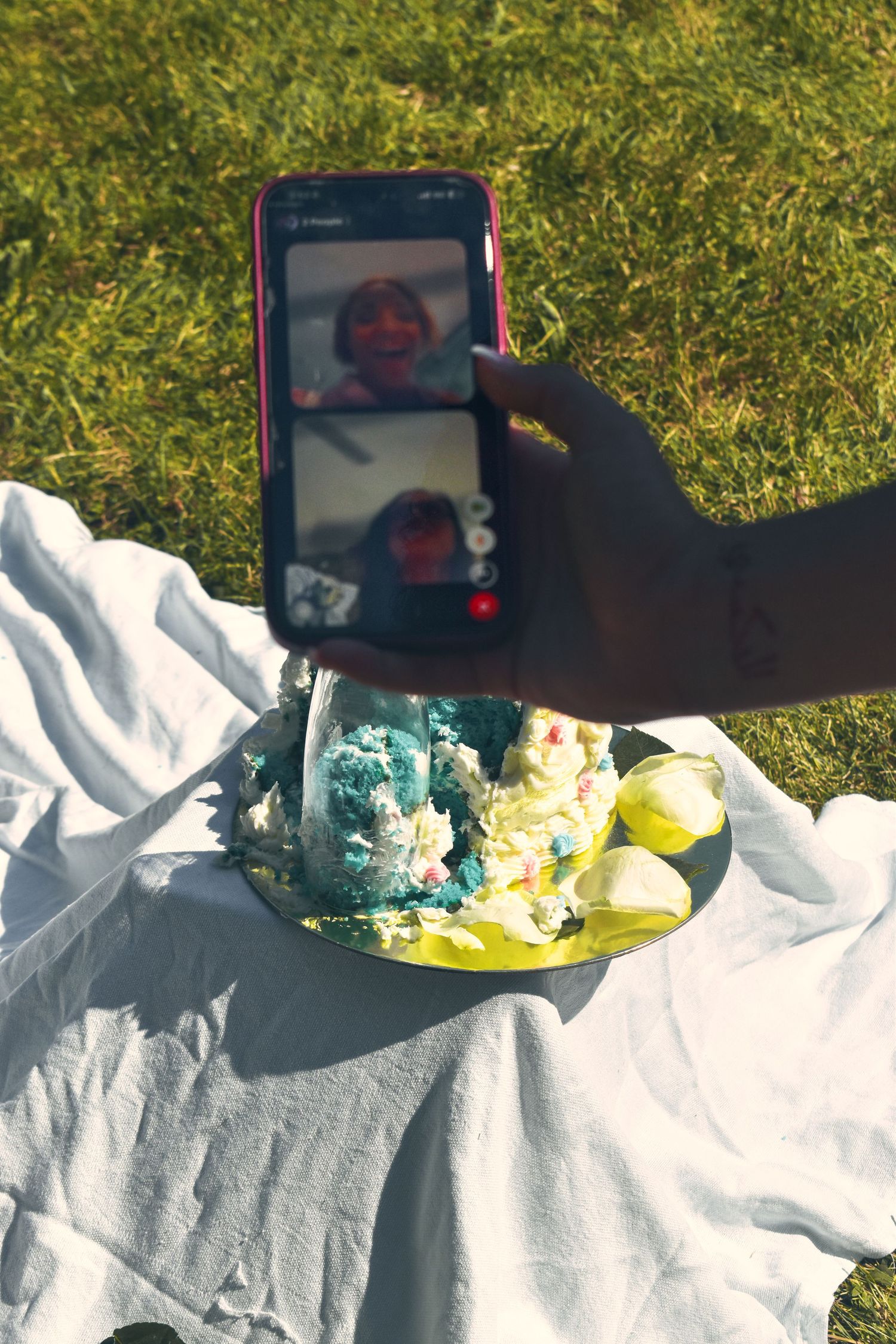 Hand holding phone on FaceTime call next to smashed birthday cake on yellow plate on white blanket.