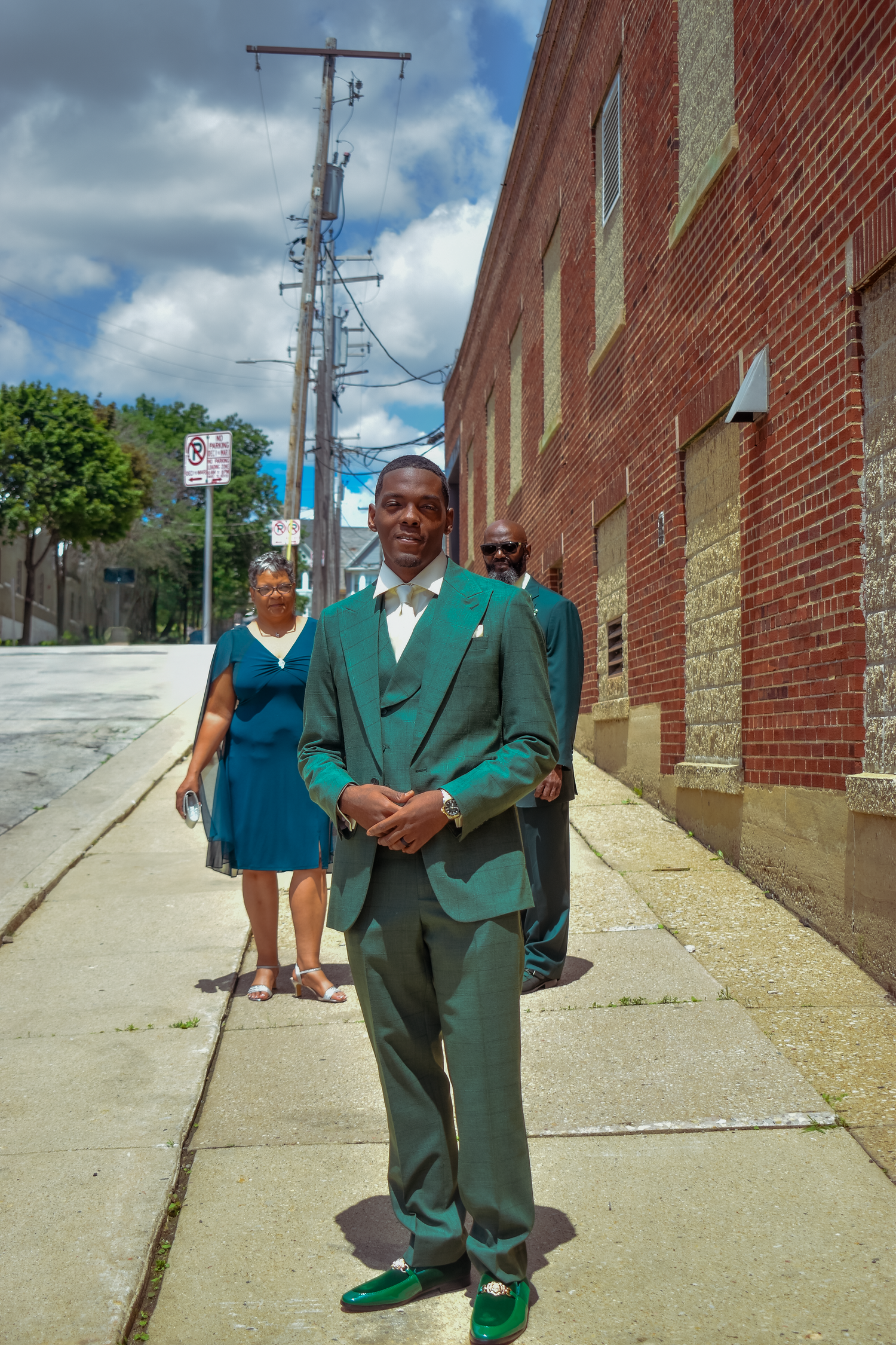 Man in green suit and shoes stands on urban sidewalk near brick building, woman in blue dress visible behind him.