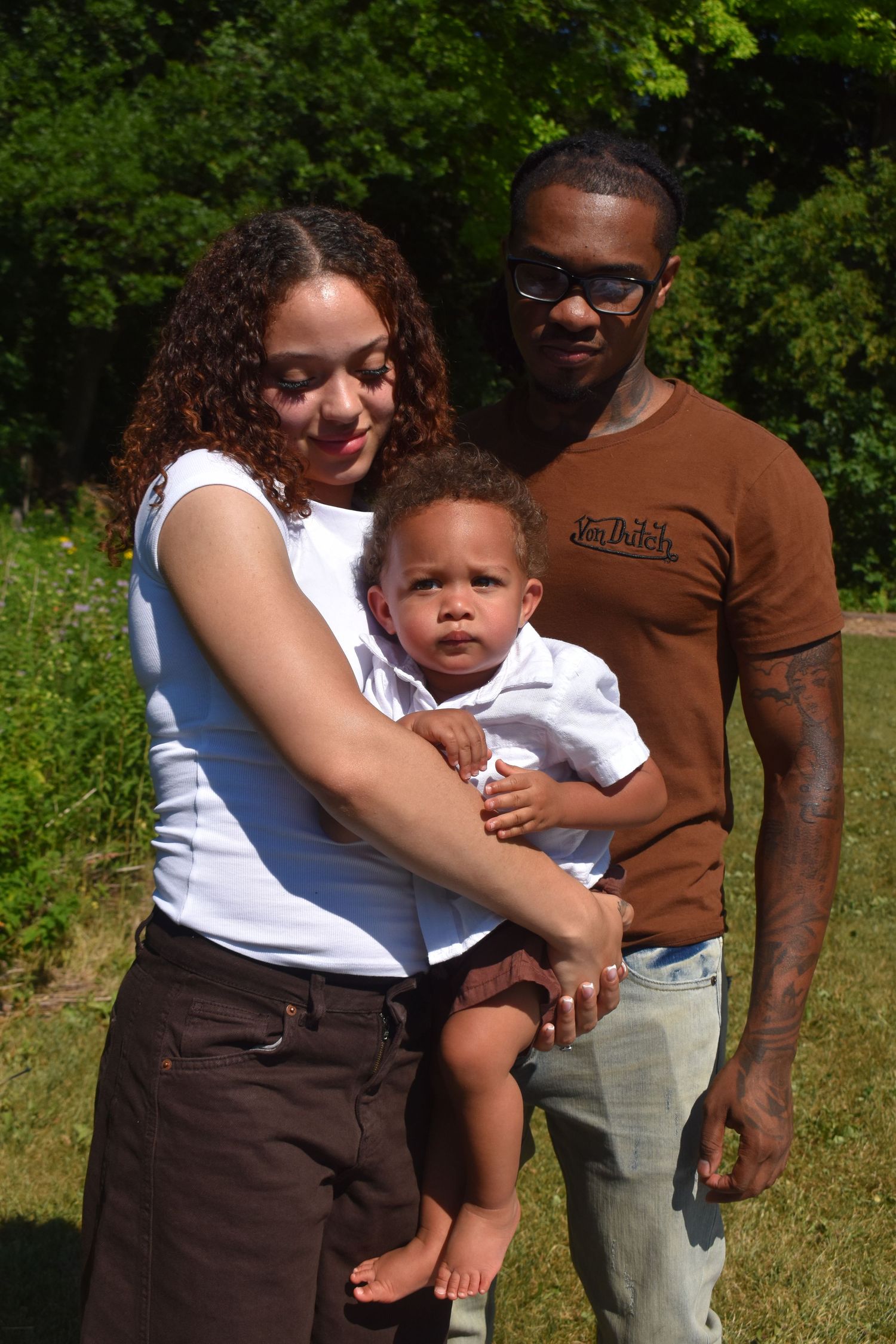 Young family posing outdoors, mother holding baby while father stands beside them in a sunny green park.