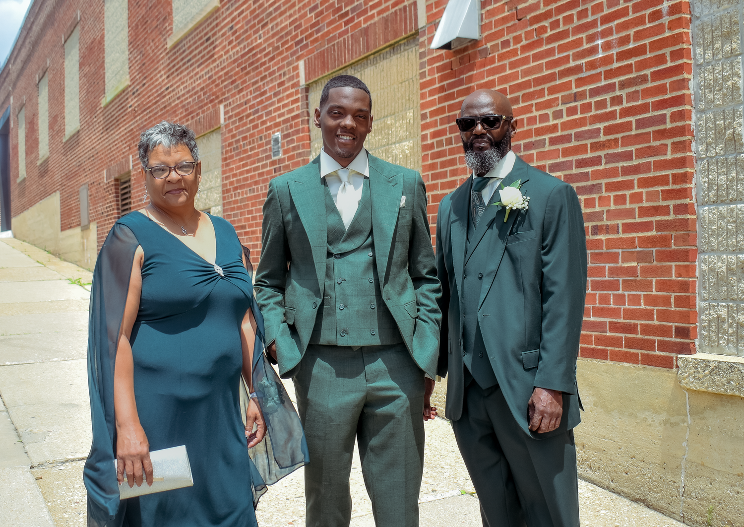 Three people dressed in elegant green formal attire pose together outside a brick building on a sunny day.