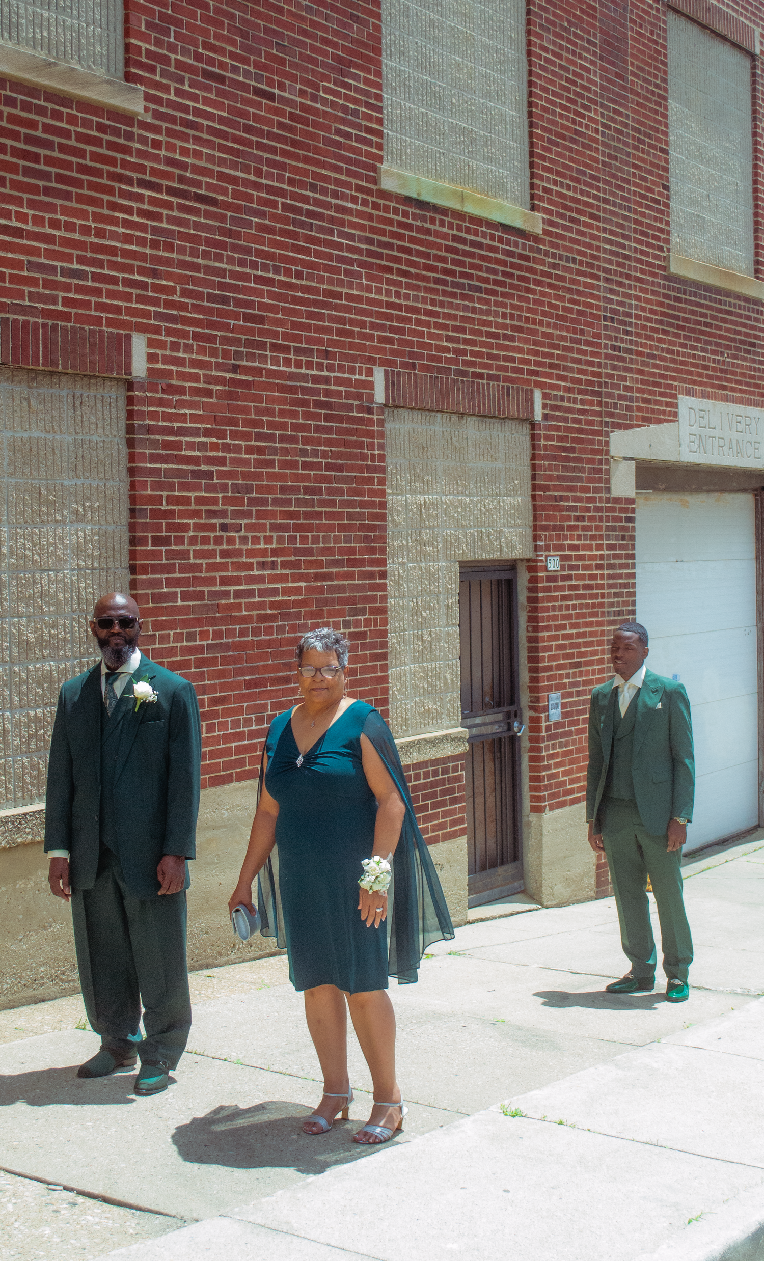 Two men in teal suits and woman in teal dress posing outside brick building on sunny day. mom and dad first look at son on wedding day.