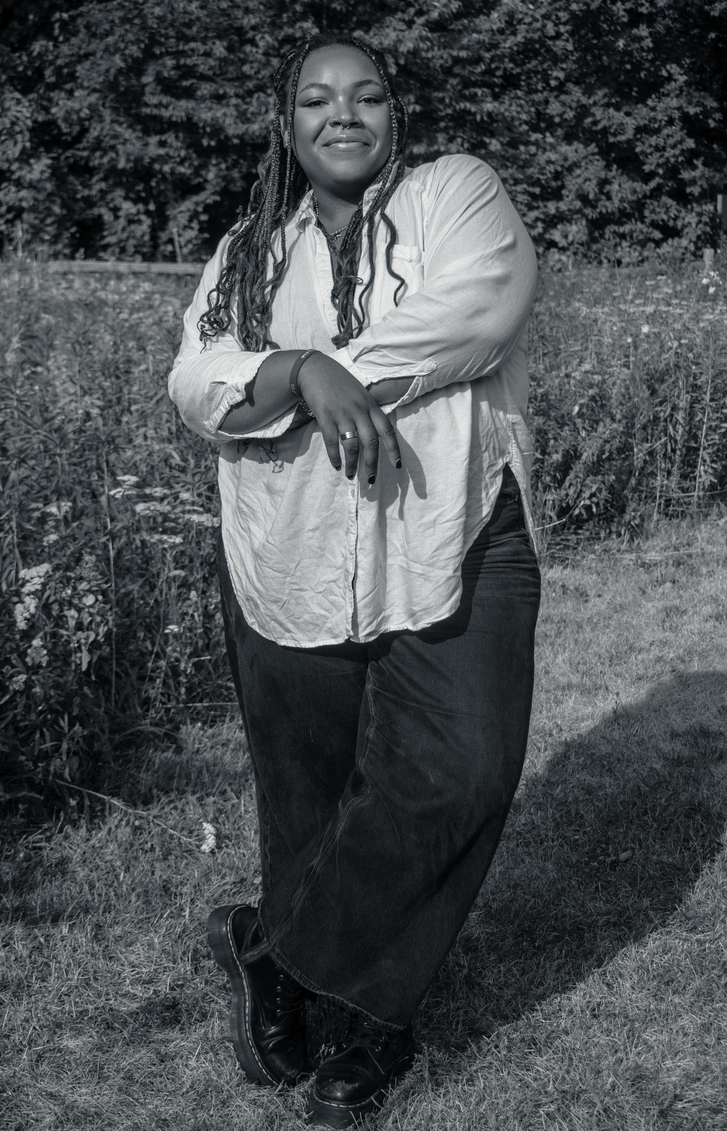 Black and white portrait of a smiling woman with long braids wearing a white shirt and wide-leg jeans outdoors.