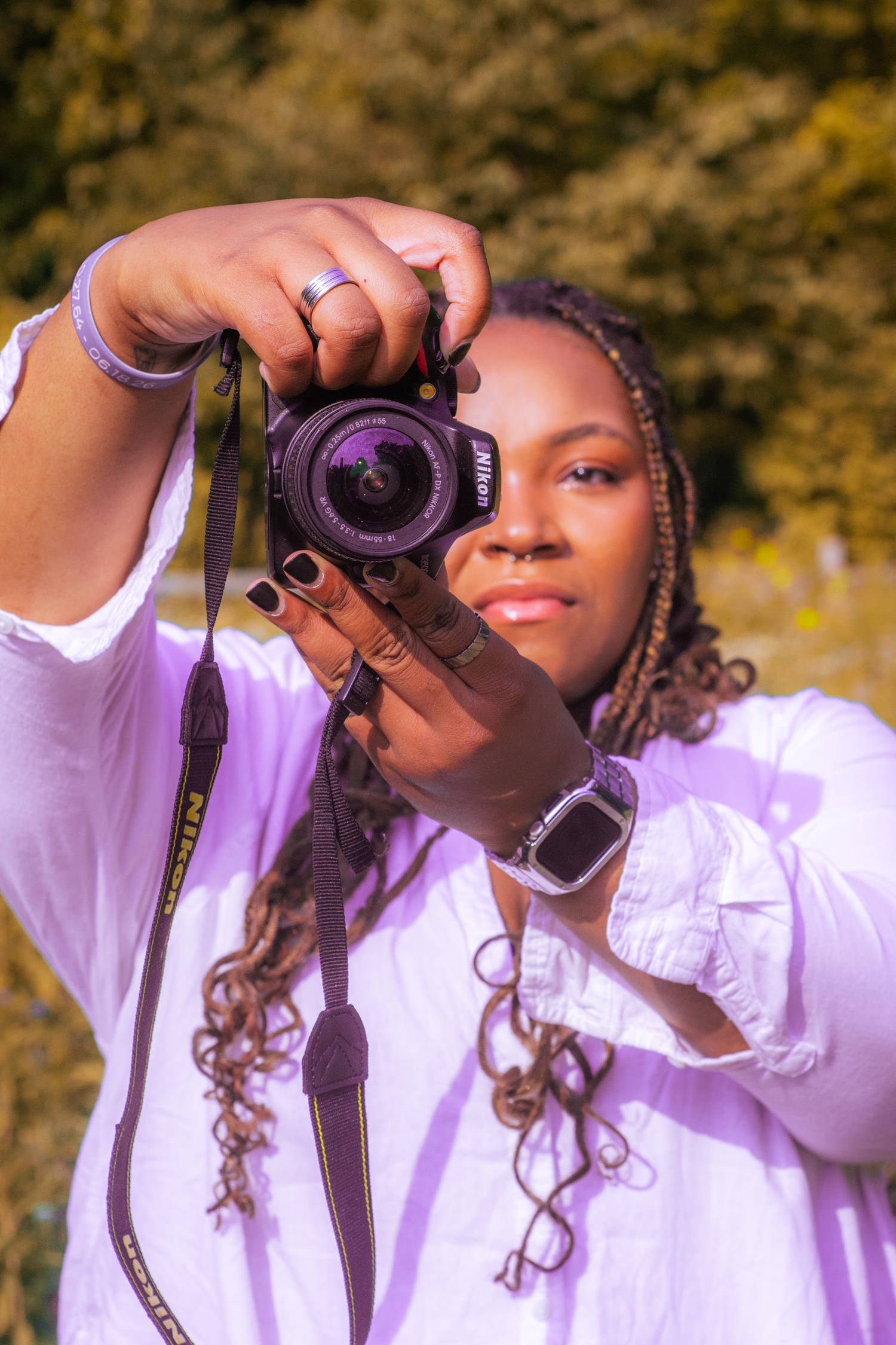 Young woman with braids holding a Nikon camera up to photograph in a sunny outdoor setting.