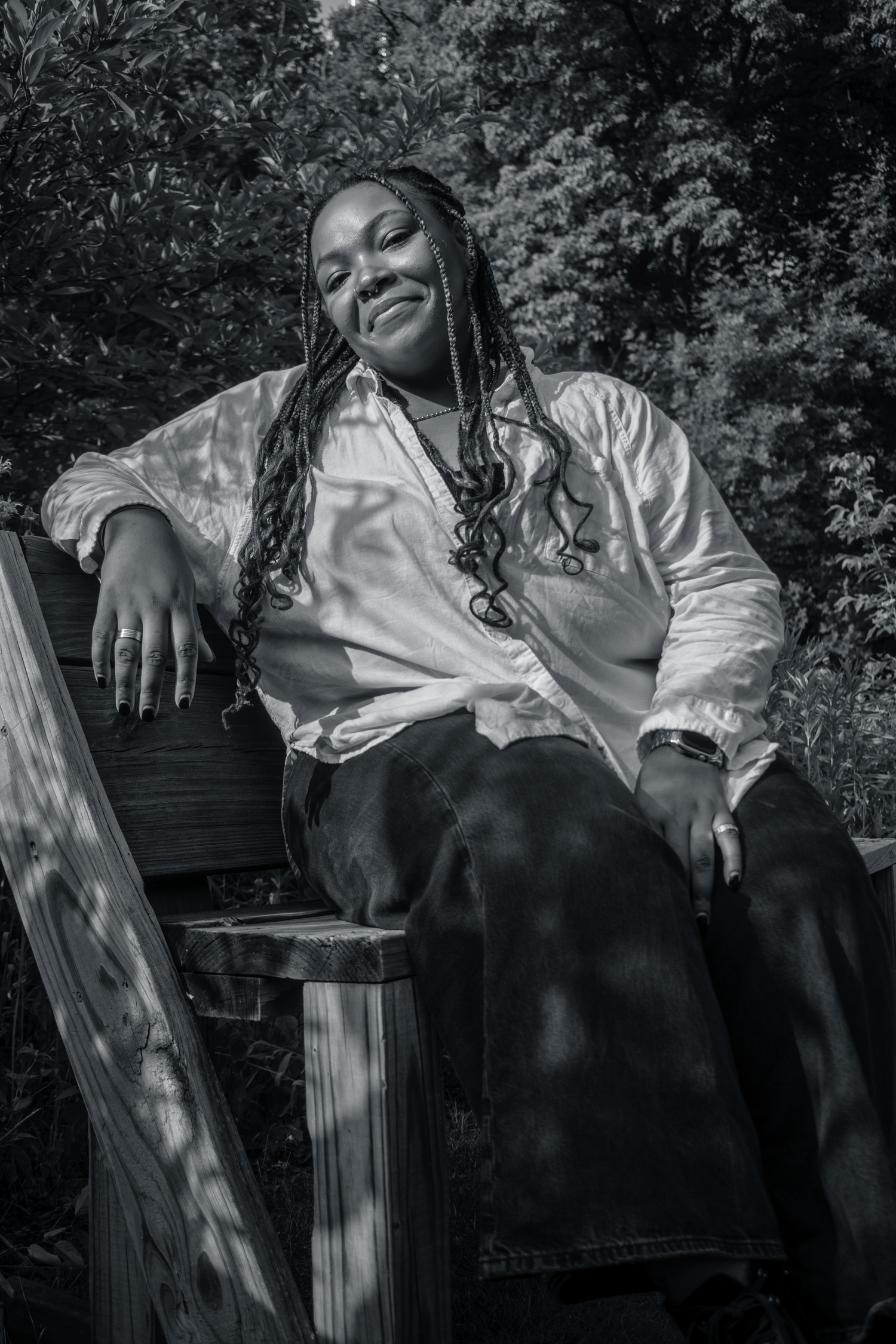 Black woman with braids reclines relaxed on wooden bench in garden, wearing white shirt and dark pants, smiling softly.