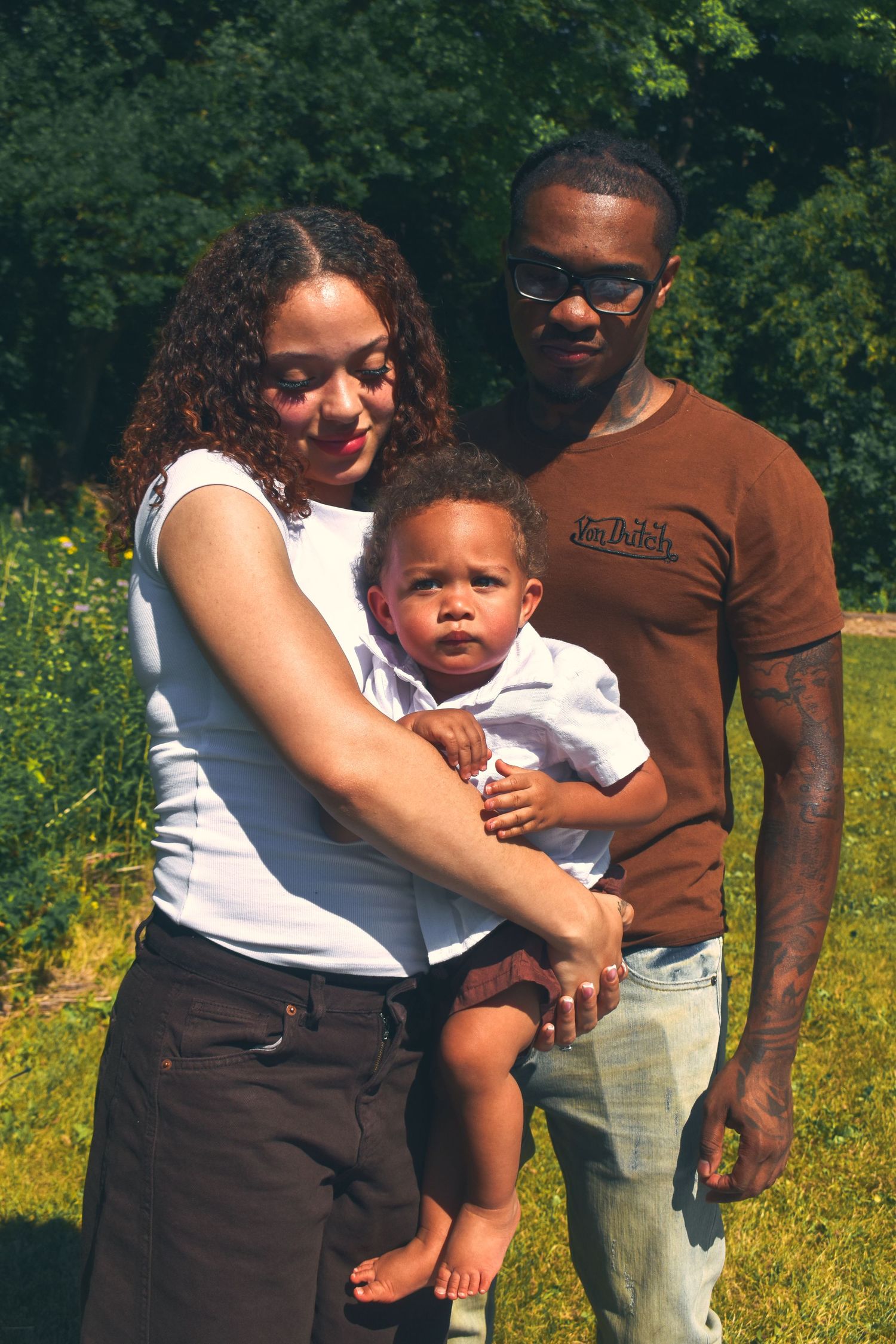 A young family poses in a sunny green field, with a mother and father holding their young toddler son.
