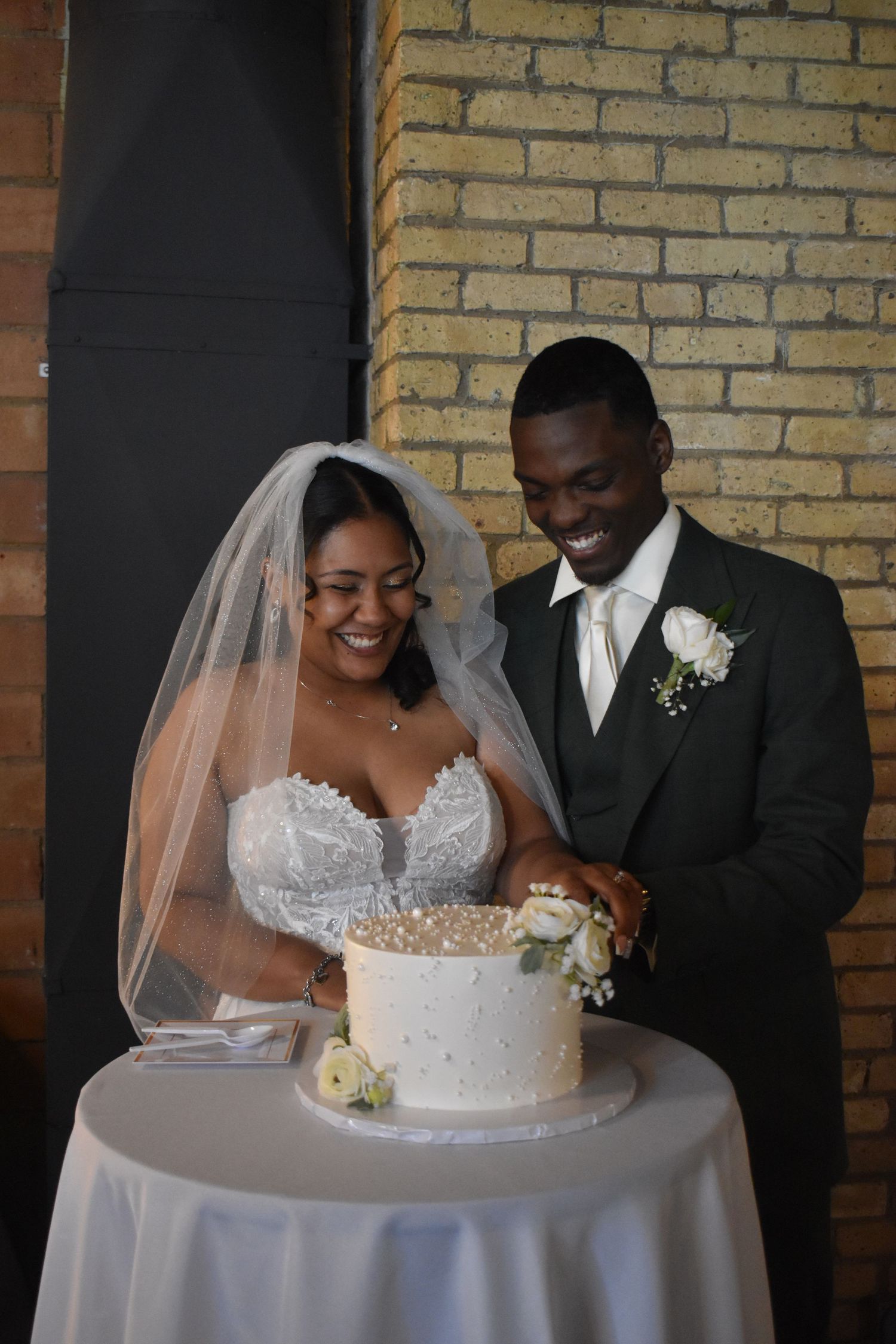 Smiling bride and groom cutting their white wedding cake together at their reception in front of a brick wall.