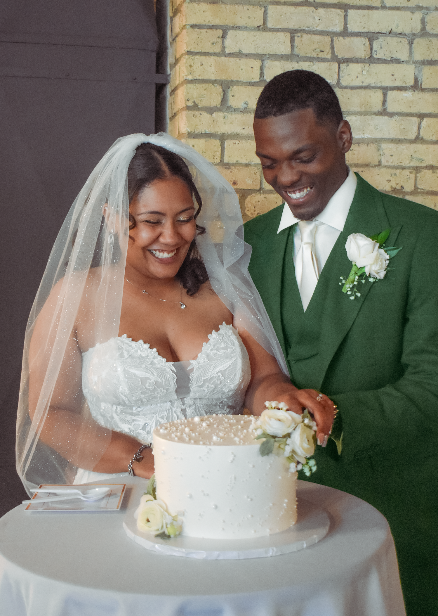 Bride and groom smiling while cutting white wedding cake decorated with pearls and flowers indoors.