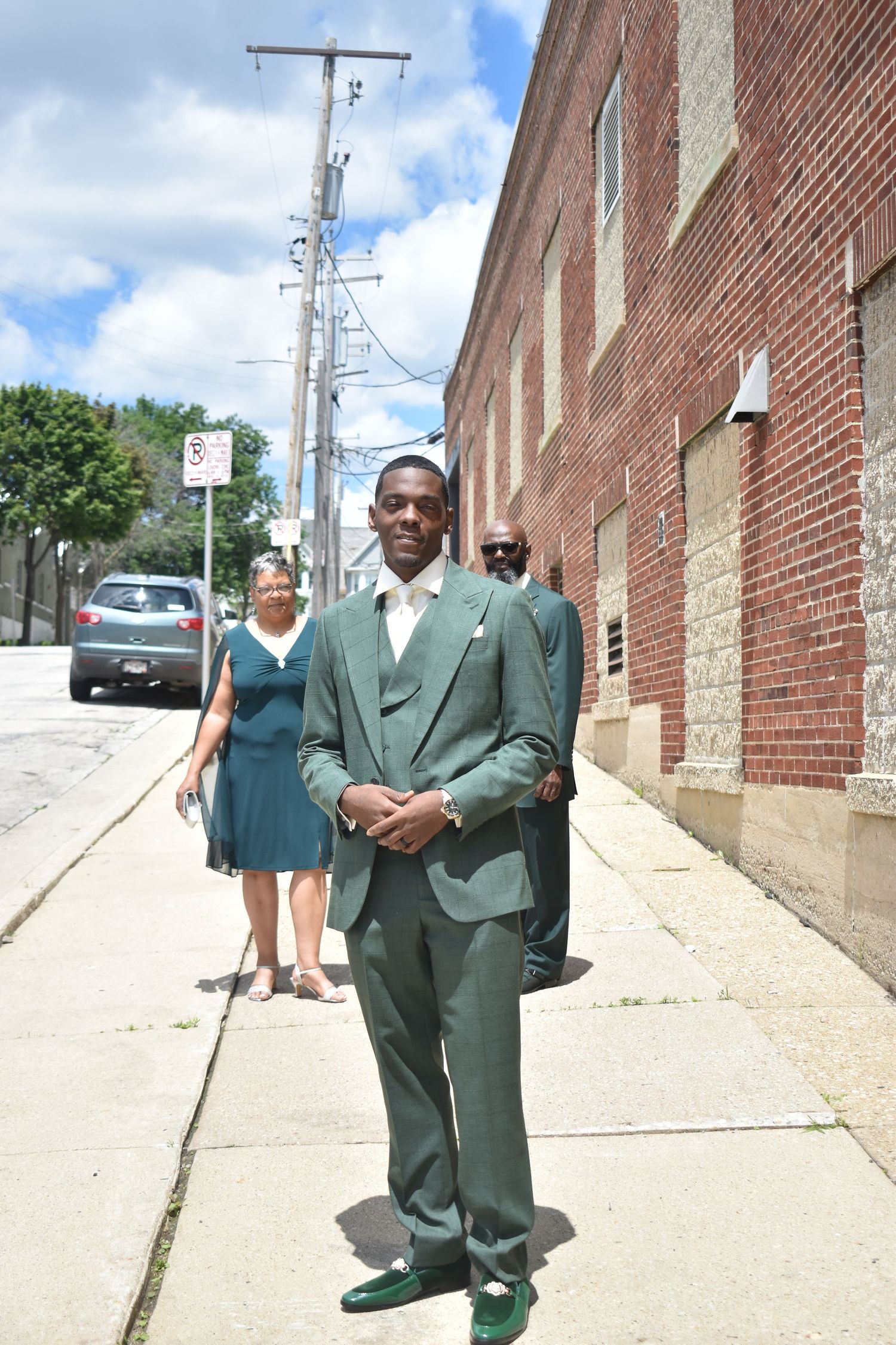 Man in green suit stands on sidewalk near brick building, with woman in teal dress and another person visible behind him.