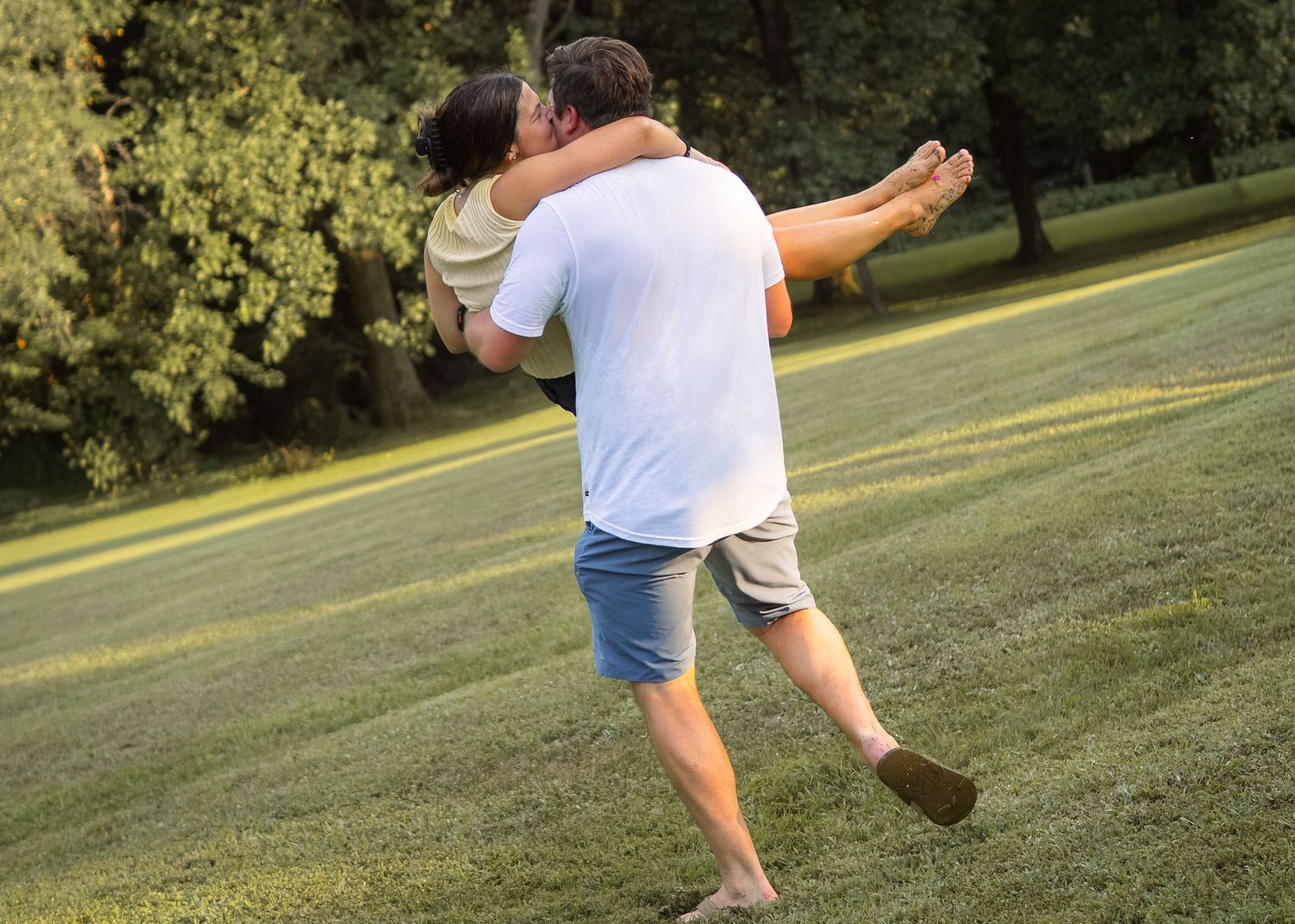 Man spins woman around in his arms while walking across lush green lawn outdoors.