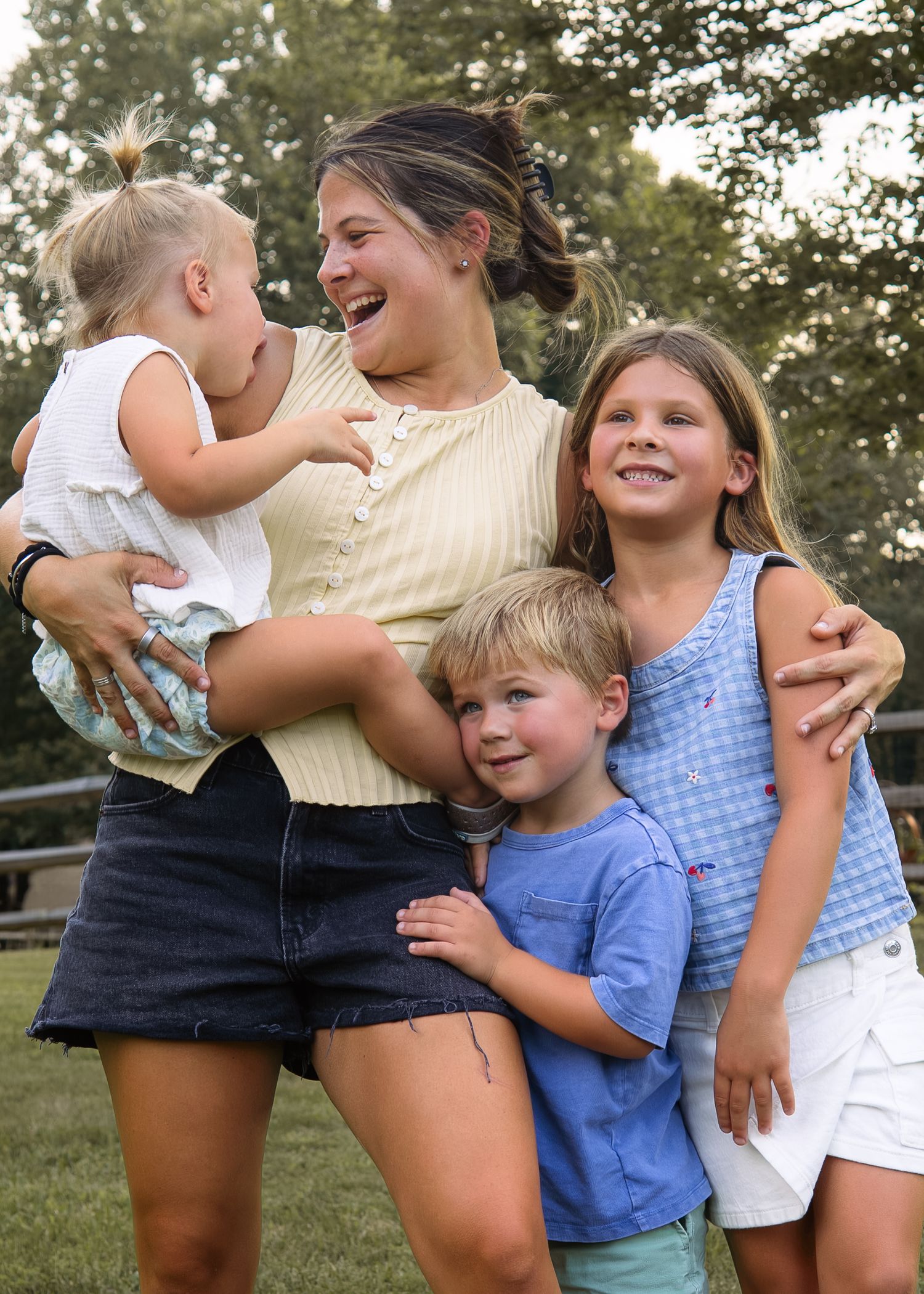 A young woman laughs while holding a toddler, hugged by two children outdoors in a green park setting.