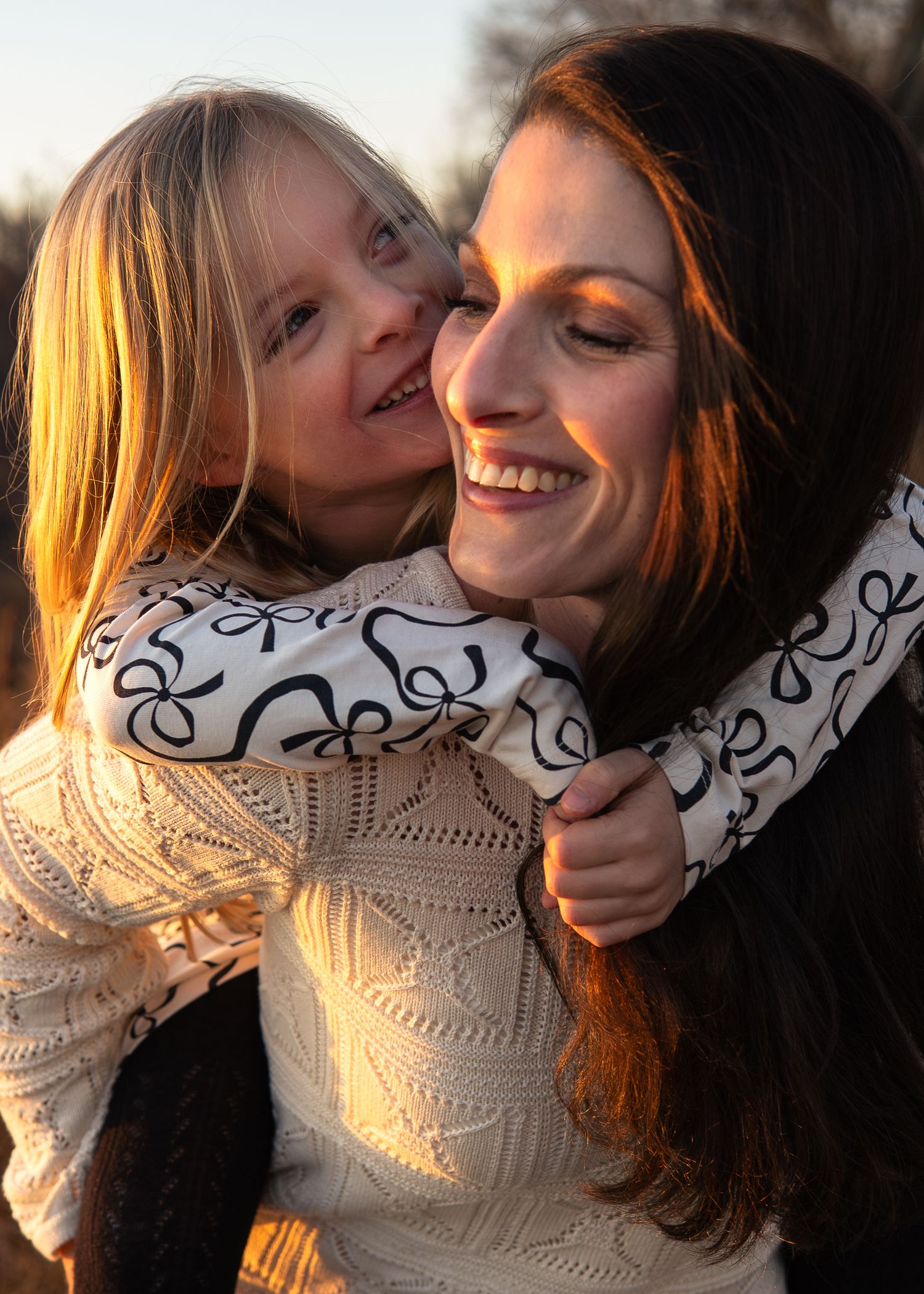 A smiling woman carries a happy young blonde girl on her back in warm golden sunset light outdoors.