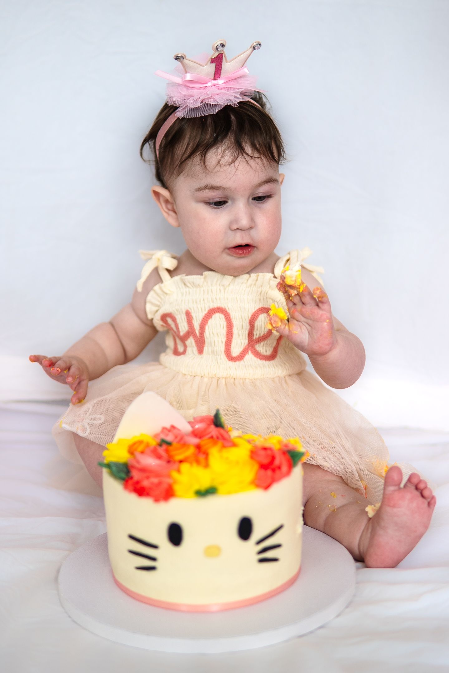 Adorable baby in cream dress and pink crown sits with a decorative Hello Kitty themed birthday cake with colorful flowers.