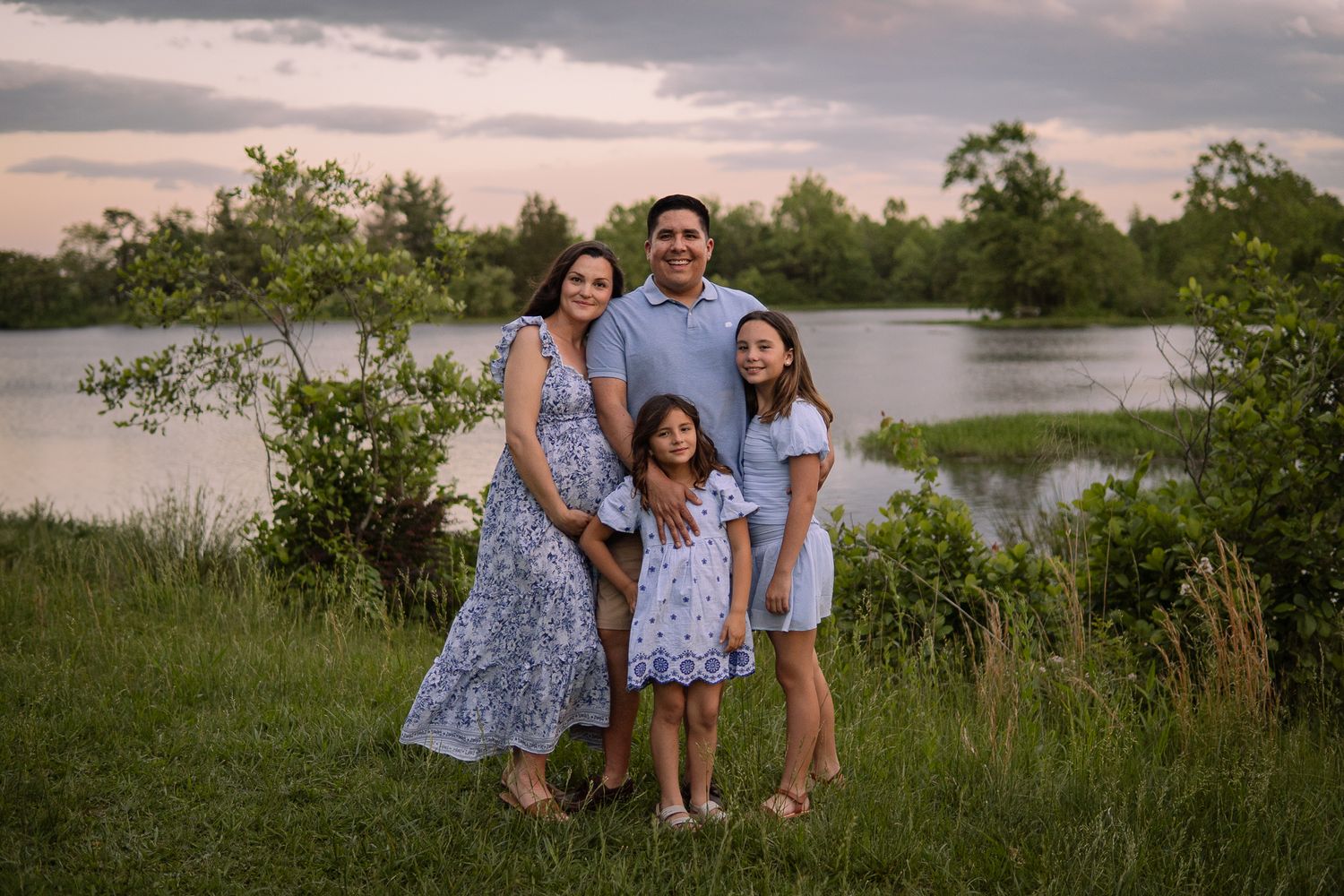 A family of four poses together by a lake during sunset in coordinated blue and white outfits.