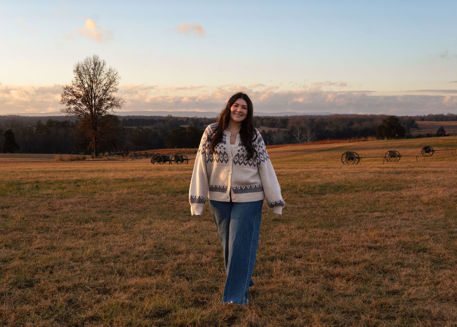 Young woman wearing a patterned white sweater and jeans posing in an open field during golden hour sunset.