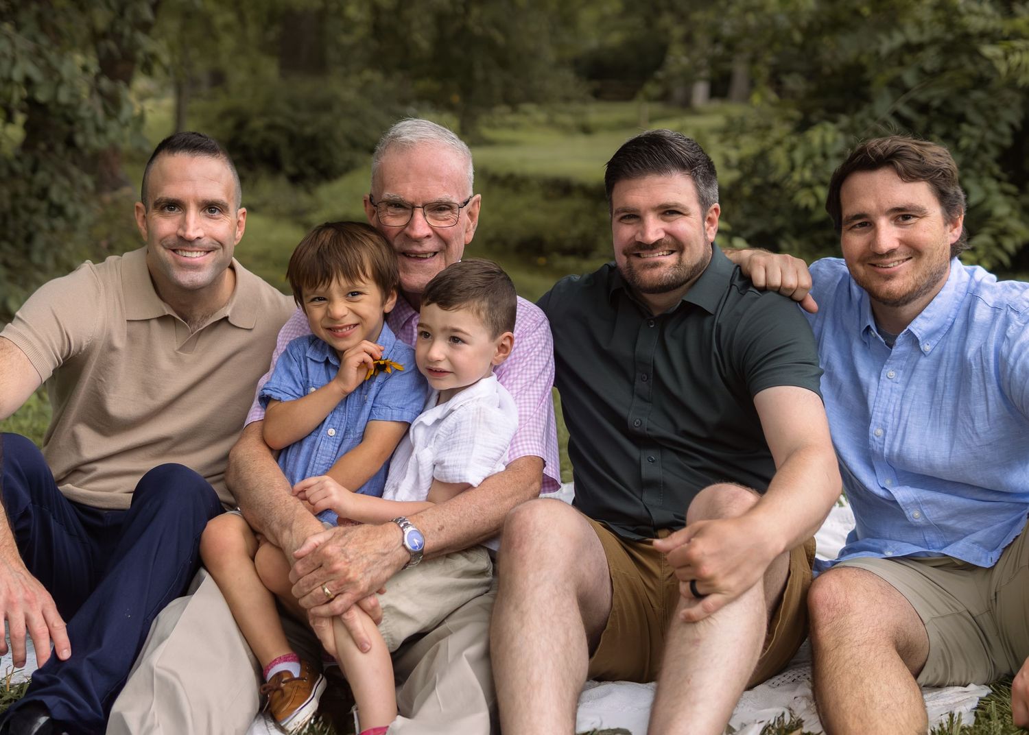 Multi-generational family portrait of five men and two young boys smiling outdoors in a lush green park setting.