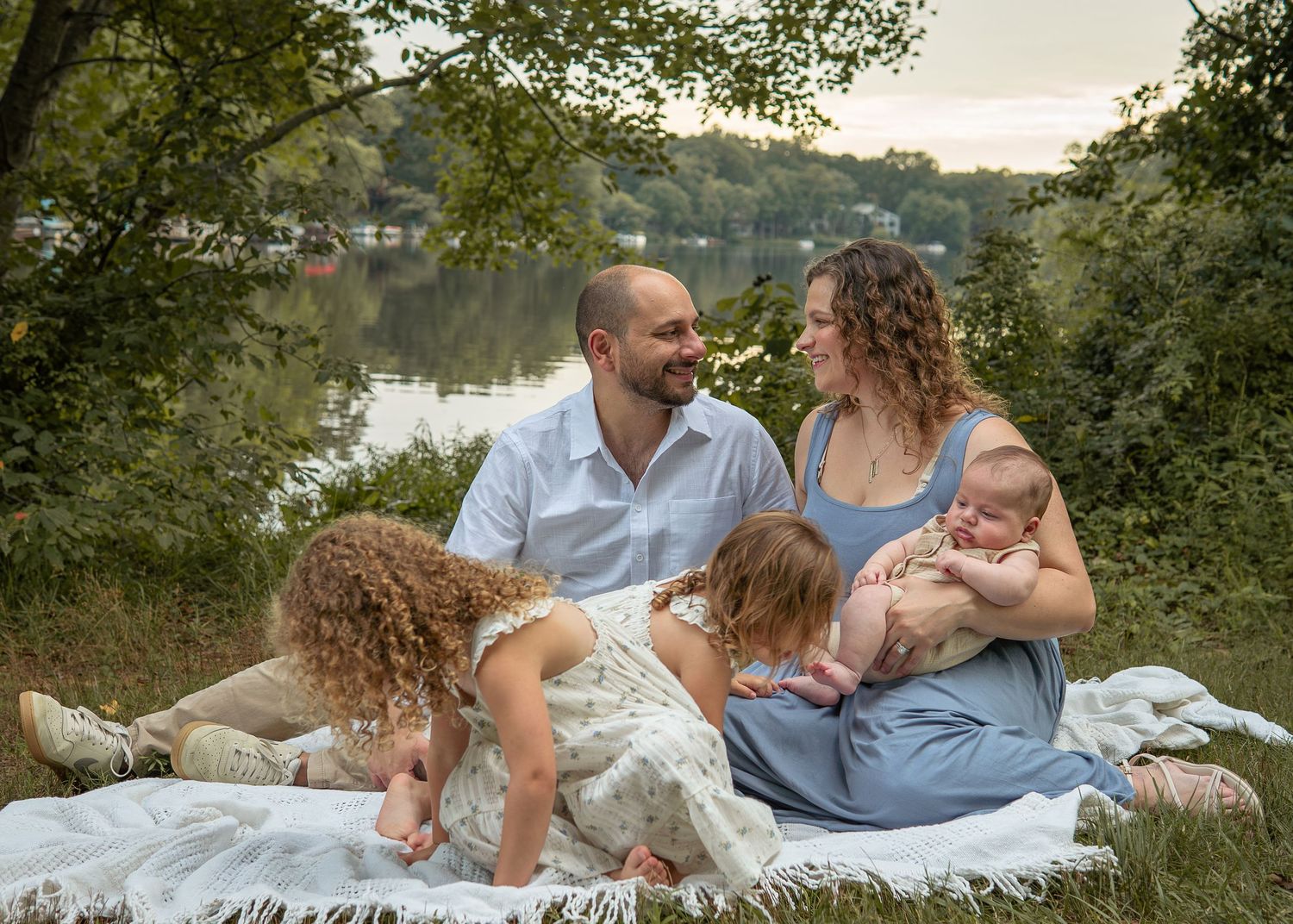 Family of four sitting on a blanket by a river, parents smiling with two young children outdoors.