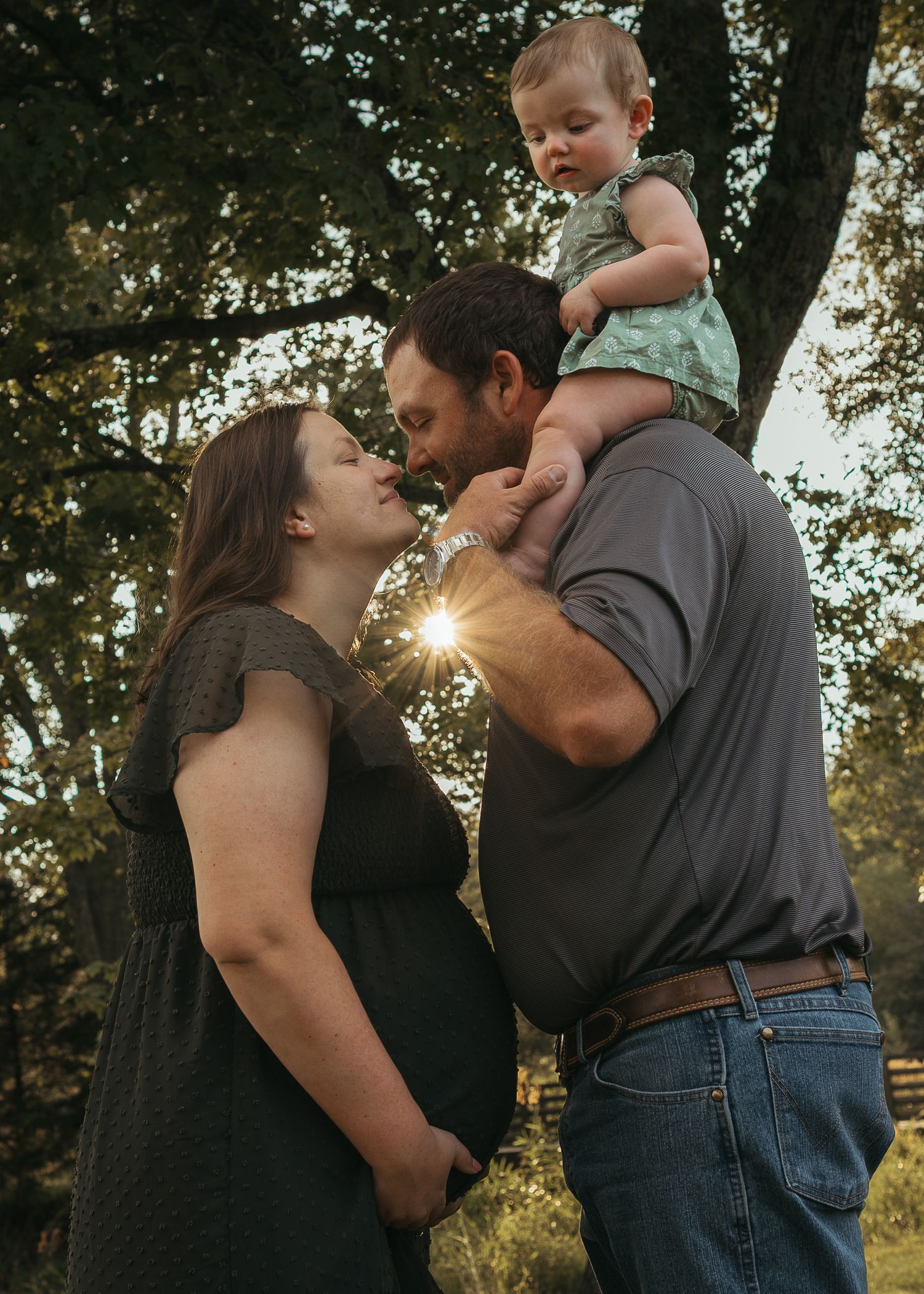 Pregnant woman and man kissing outdoors at golden hour while holding a baby in a green onesie among trees.