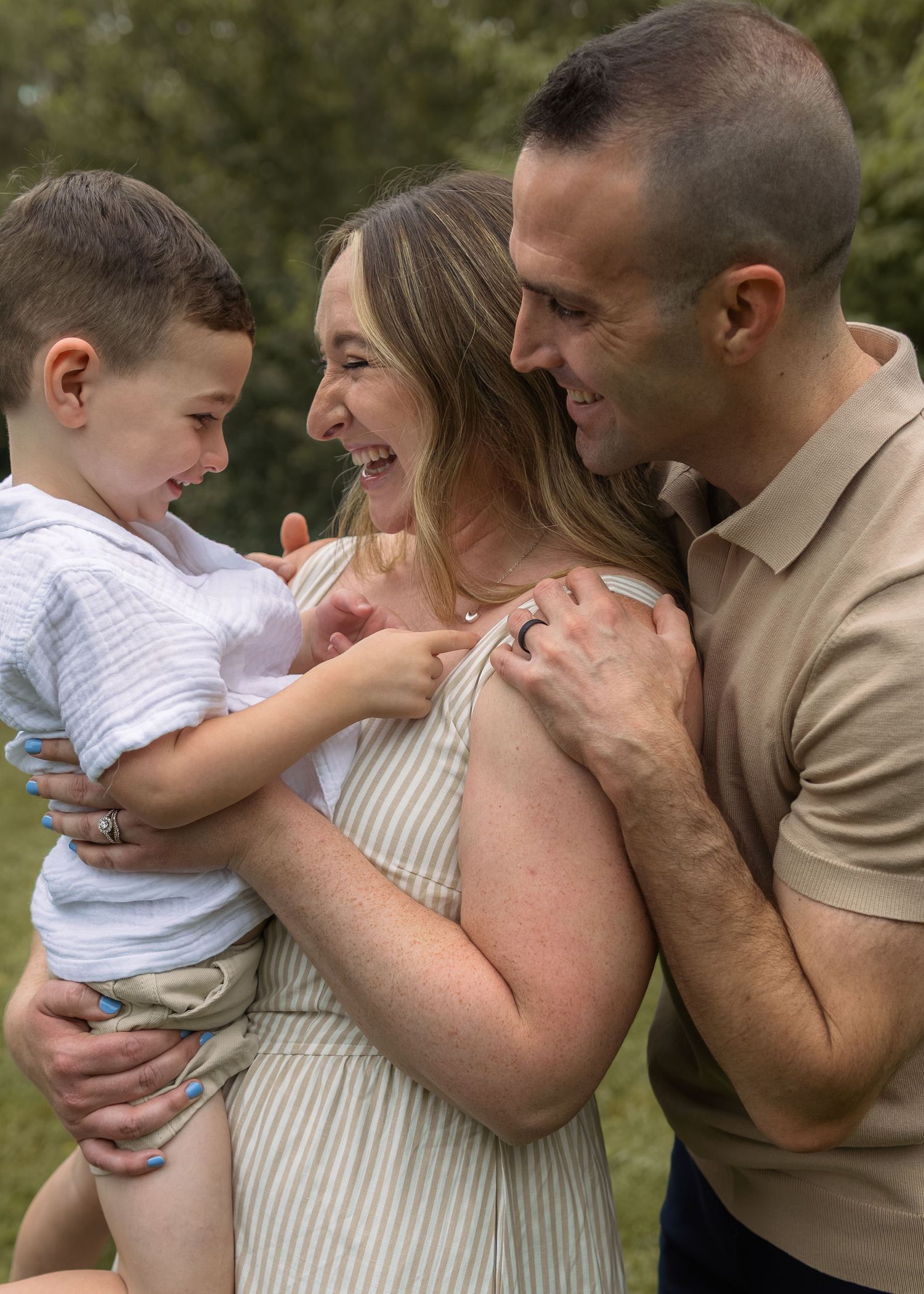 Happy couple laughing together while holding their young toddler son outdoors in a lush green park.