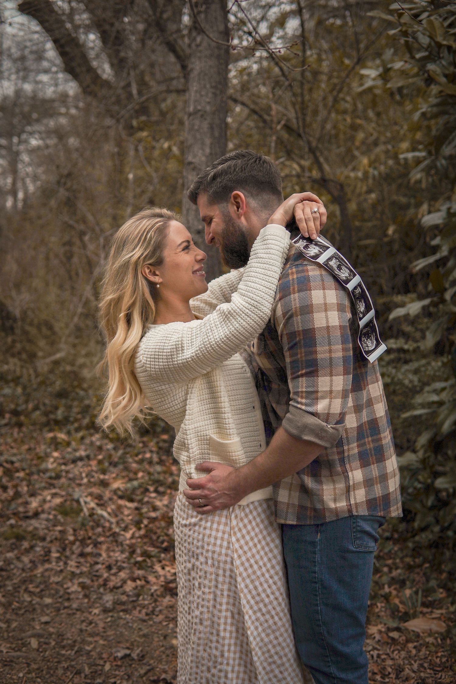 A romantic autumn outdoor portrait of a couple embracing in casual sweaters among fallen leaves and trees.