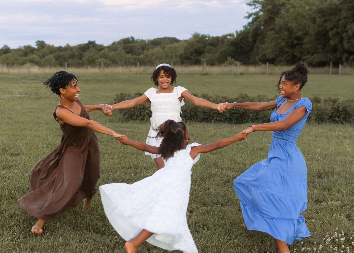 Four women joyfully spin in a circle holding hands in a lush green meadow on a cloudy day.