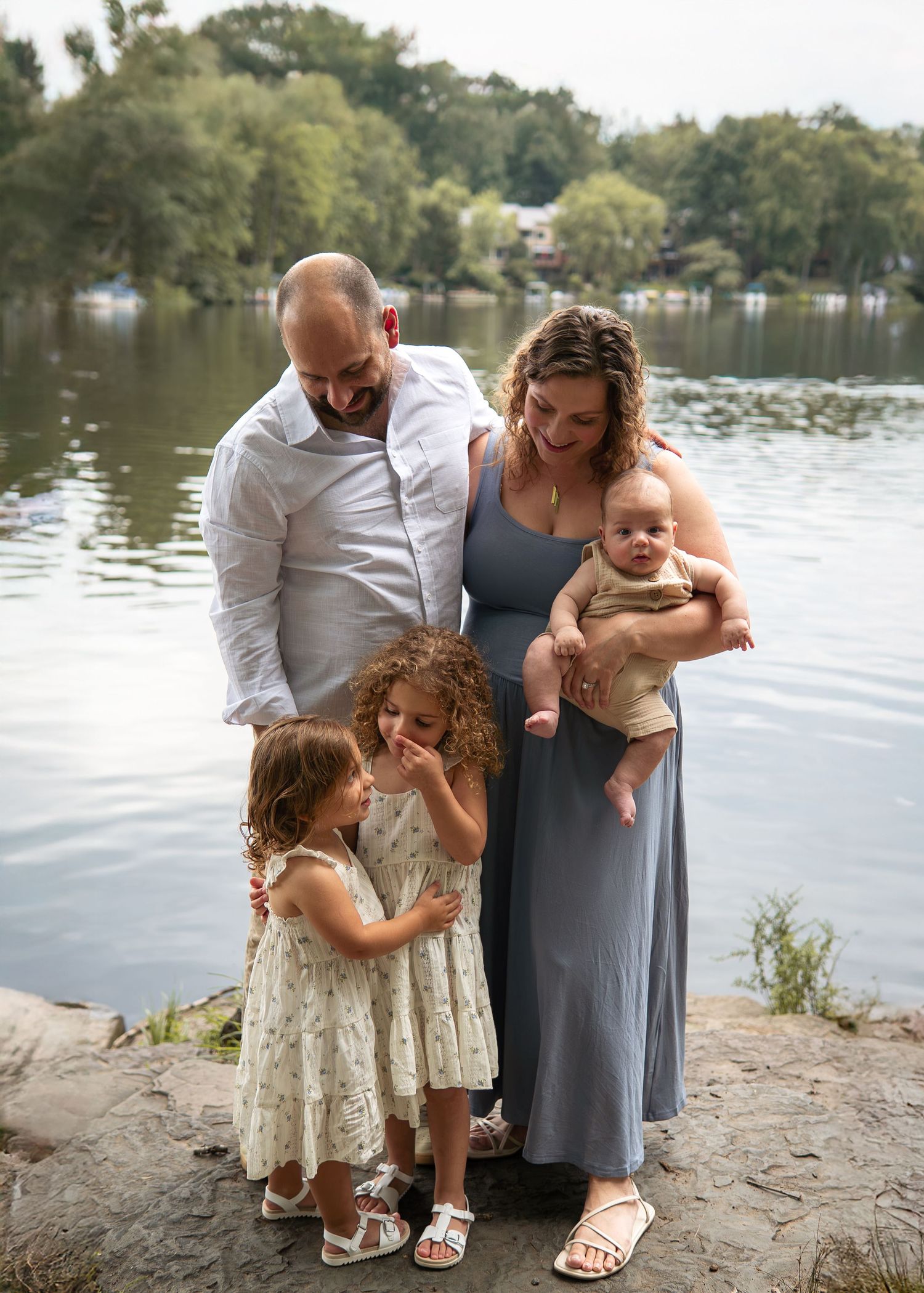 Family of five poses by a serene lake, parents smiling at baby while two curly-haired girls in floral dresses stand close.