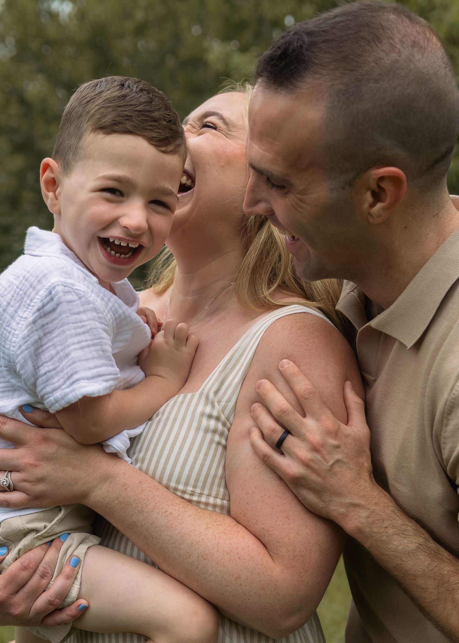 A joyful family of three laughing outdoors, with parents holding their young son in a warm, candid moment.